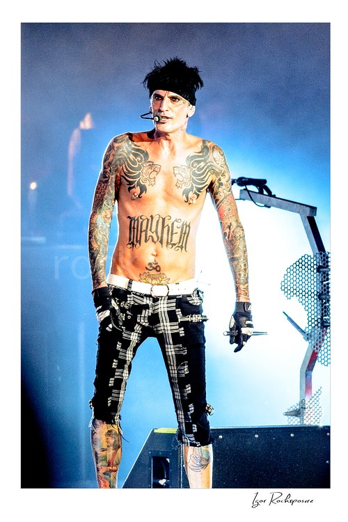 Vertical color image of Tommy Lee performing live during a Mötley Crüe concert, shirtless with tattoos and blue stage light