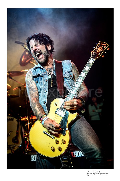 Vertical color image of Tracii Guns performing live with electric guitar amid stage smoke