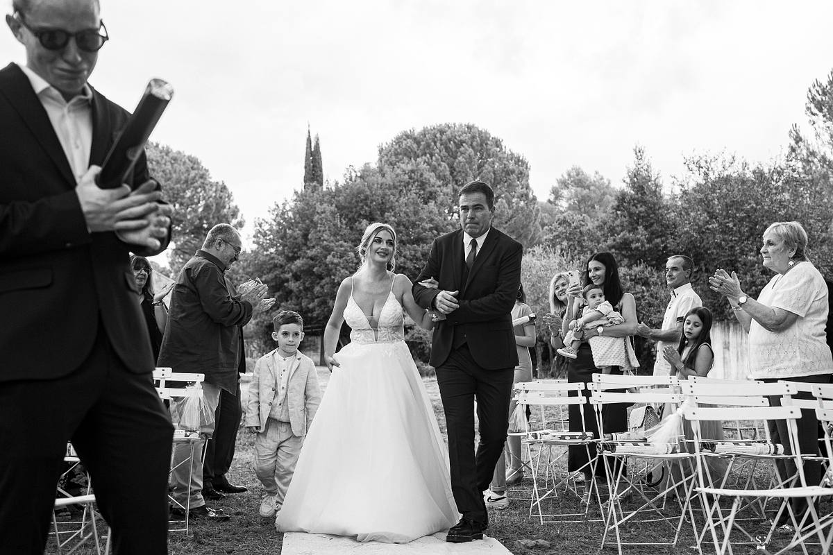 La mari&eacute;e rayonnante, accompagn&eacute;e de son p&egrave;re, passe devant des invit&eacute;s applaudissant sur des chaises blanches dans la prairie, avec le mari&eacute; qui attend avec impatience, illustrant l'all&eacute;e menant au 'oui' sous le ciel de N&icirc;me
