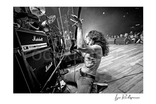 Horizontal black and white image of Brent Woods playing guitar shirtless on the stage floor beside Marshall amplifier stacks with the crowd visible beyond