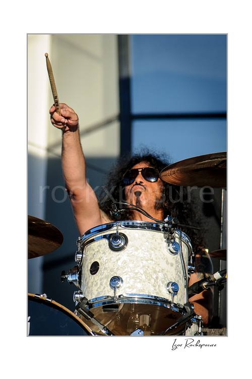 Vertical color image of Frankie Banali behind a white drum kit with one drumstick raised high during a live performance