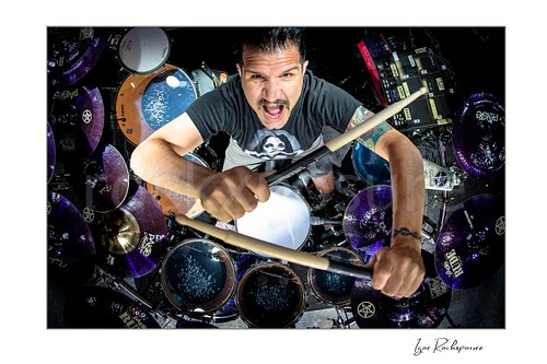 Horizontal color aerial image of Charlie Benante of Anthrax performing live on stage behind his drum kit, photographed from above with drumsticks extended toward the camera and the full set visible below