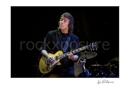 Horizontal color image of Steve Hackett performing live with electric guitar on stage with the drum kit behind him