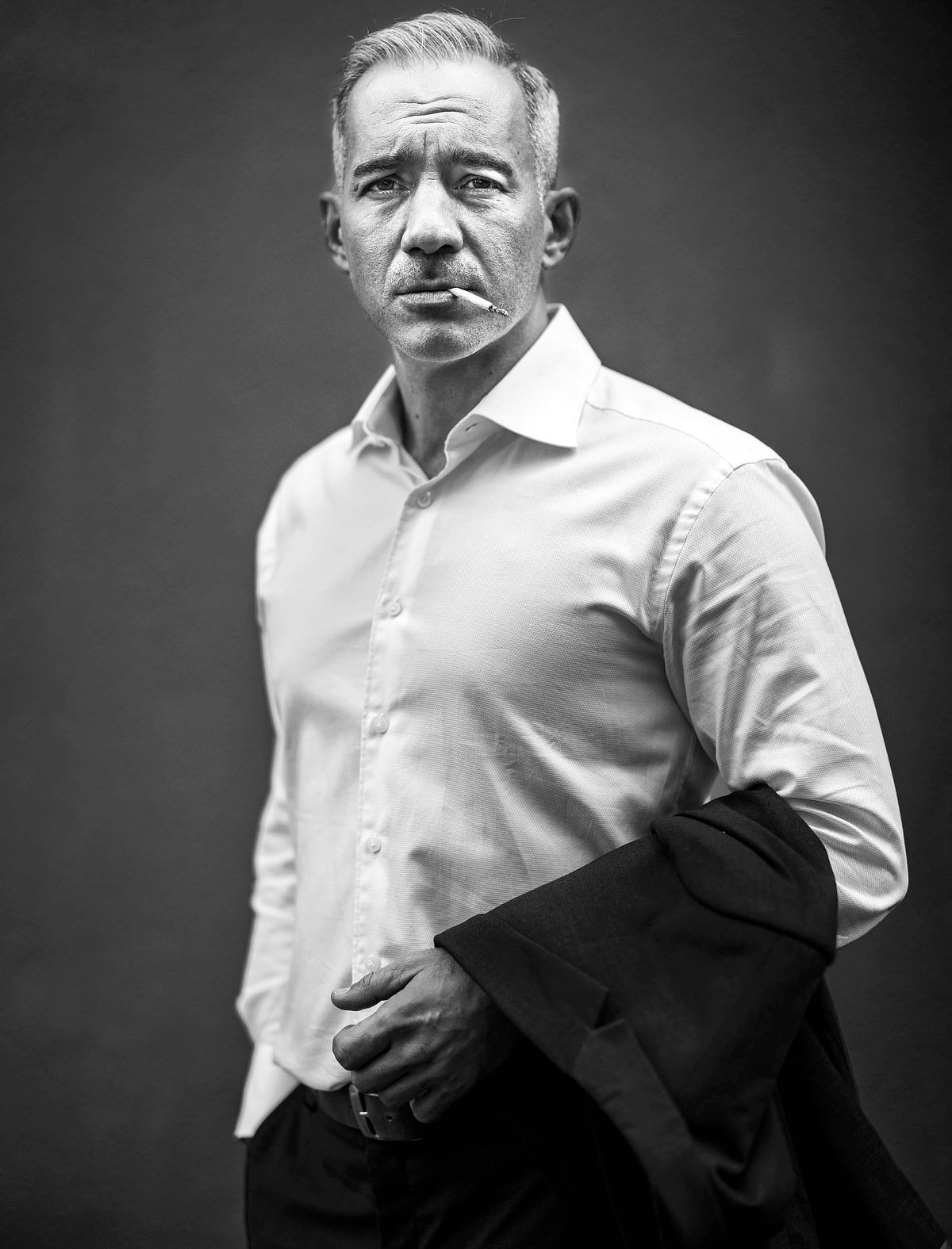 Portrait of a man wearing a white short with his suit draped over his arm. He smokes a ciggarette.