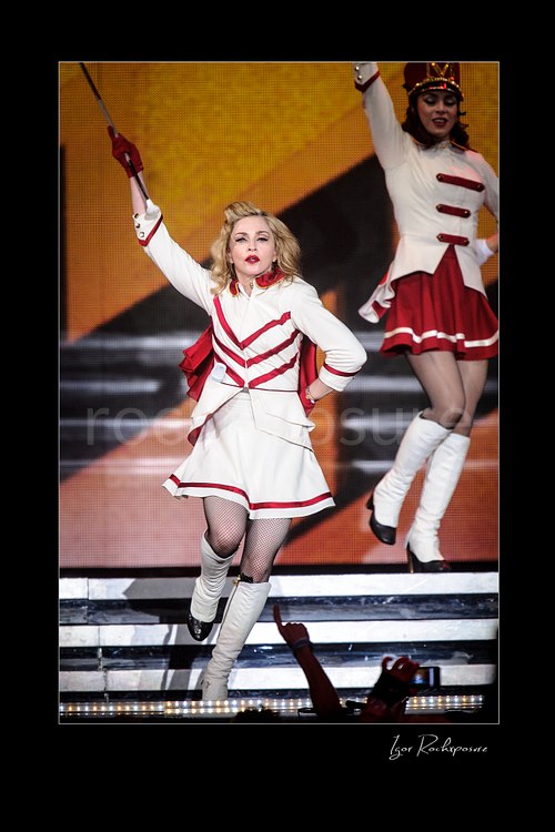 Vertical color image of Madonna performing onstage in a white and red stage outfit with one arm raised while a dancer appears behind her
