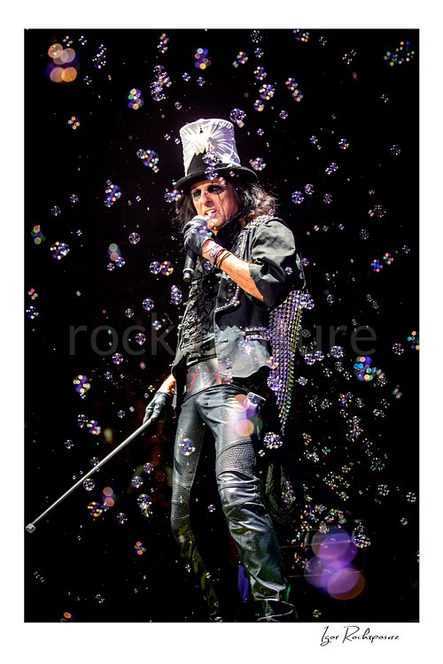 Vertical color image of Alice Cooper performing live on stage in a silver top hat with a microphone, cane, black leather outfit, and bubbles floating around him