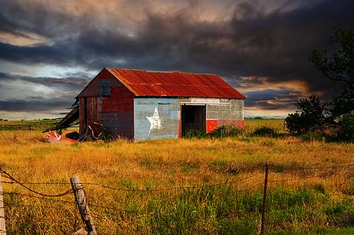 Weathered metal barn painted with the Texas state flag in a golden field under a dramatic storm sky.
