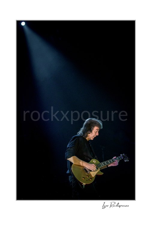 Vertical color image of Steve Hackett performing live with electric guitar beneath a single overhead spotlight