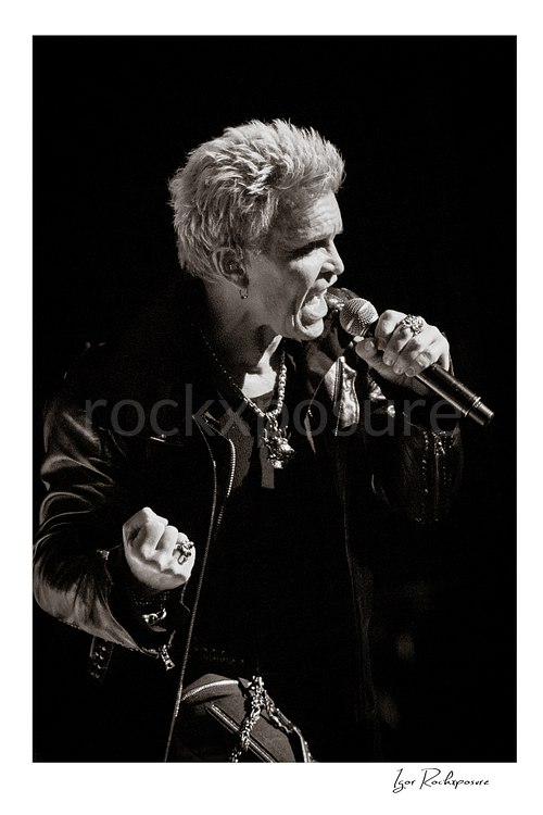 Vertical black and white image of Billy Idol singing into a microphone in a tight live performance portrait
