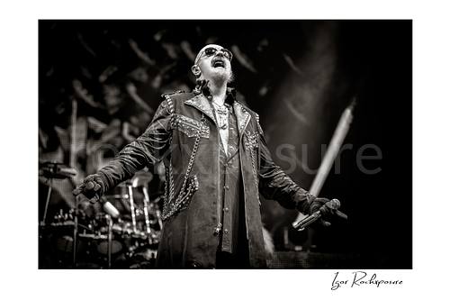 Horizontal black and white image of Rob Halford of Judas Priest performing live on stage, holding a microphone with his arms extended and the drum kit visible in the background