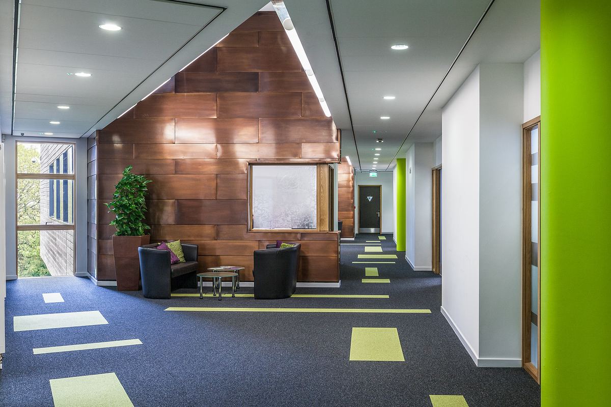 Modern office hallway with green and brown accents, seating area featuring two black chairs and a small table, and geometric carpet patterns.