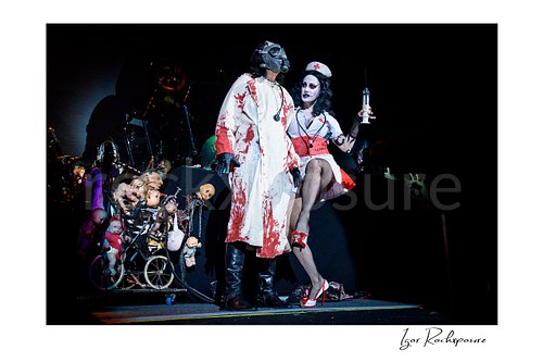 Horizontal color image of Alice Cooper performing live on stage in a blood-stained straitjacket beside his wife dressed as a nurse holding a large syringe