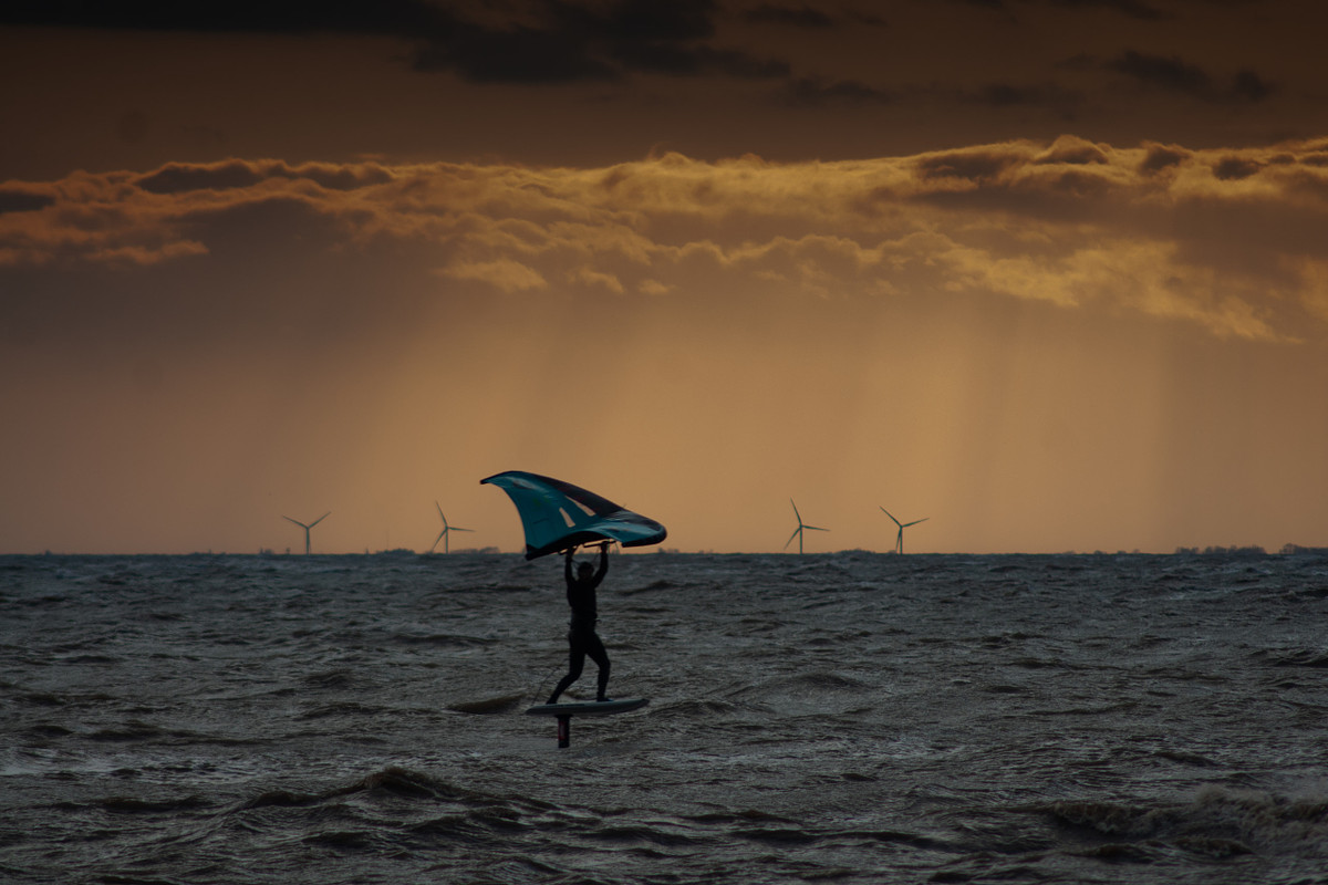 A wing foiler riding the waves off Snettisham, with offshore wind turbines on the horizon.  Alt text: