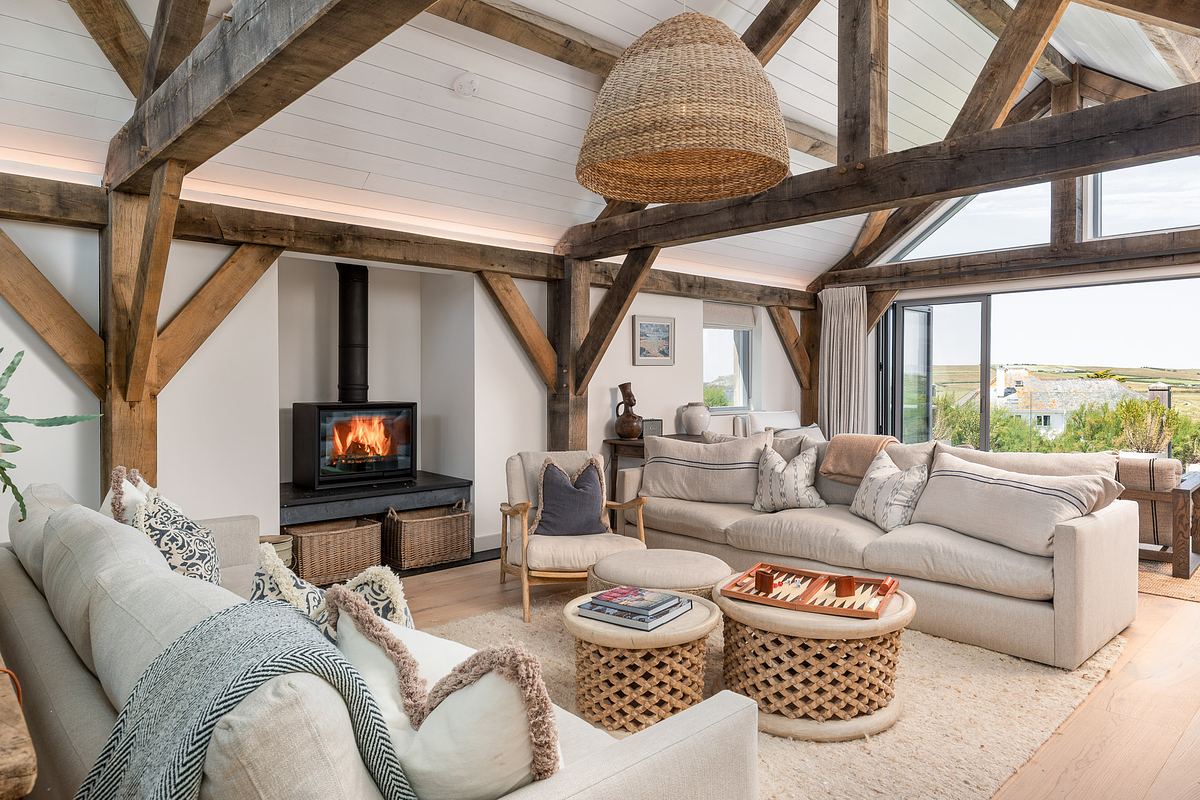 Modern rustic living room with exposed wooden beams, light sofas, a wood-burning stove, woven decor, coffee tables, and large windows overlooking a landscape.