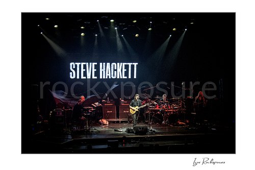 Horizontal color image of Steve Hackett performing on stage with his full band beneath spotlights and a large backdrop screen