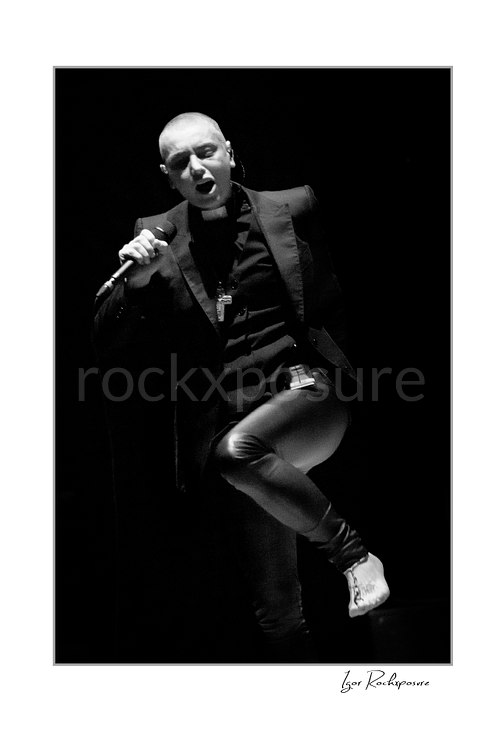 Sinéad O’Connor sings with her mouth open, microphone near her face and one bare foot raised across her body in a black and white stage portrait.
