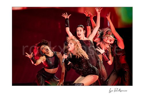 Horizontal color image of Taylor Swift singing on stage with dancers under vivid red concert lighting and dramatic motion