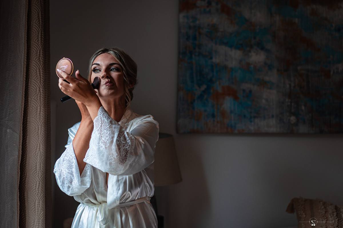 La mari&eacute;e se maquille avec attention, tenant un petit miroir, &eacute;clair&eacute;e par la douce lumi&egrave;re naturelle pr&egrave;s d'une fen&ecirc;tre, avec un tableau abstrait en arri&egrave;re-plan &agrave; N&icirc;mes, immortalis&eacute;e par S&eacute;bastien Clavel