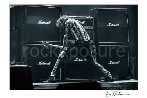 Horizontal black and white image of Nuno Bettencourt performing live on stage with an electric guitar before a wall of Marshall amplifiers