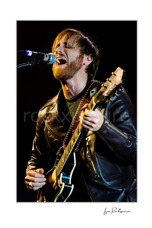 Vertical color image of Dan Auerbach of The Black Keys singing into a microphone while playing guitar in a black leather jacket
