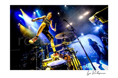 Horizontal color image of Zoltan Chaney standing on his drum kit with drumsticks under bright blue and yellow stage lights