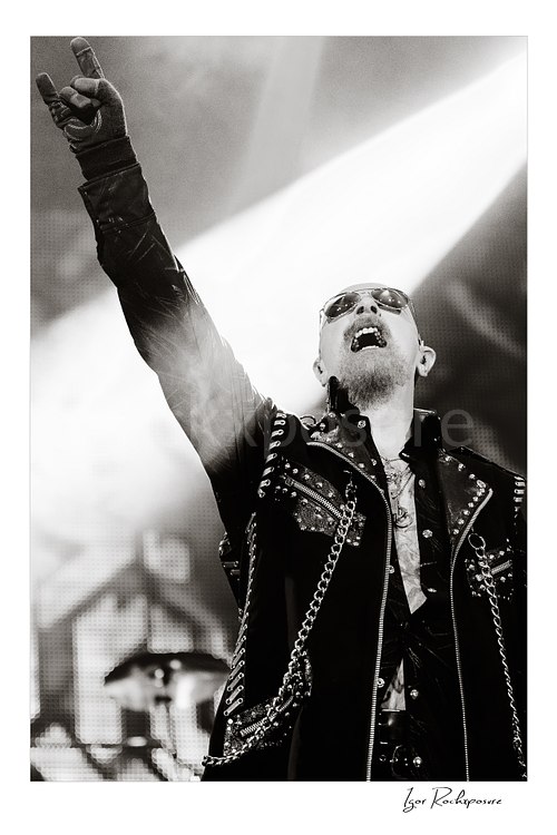 Vertical black and white image of Rob Halford of Judas Priest performing live on stage, raising one arm while holding a microphone under dramatic concert lighting