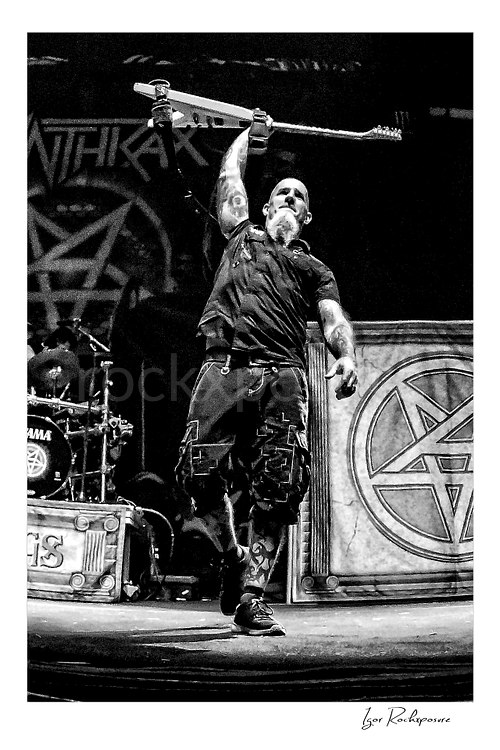 Vertical black and white image of Scott Ian of Anthrax performing live on stage, raising his guitar overhead with the band’s stage set visible in the background
