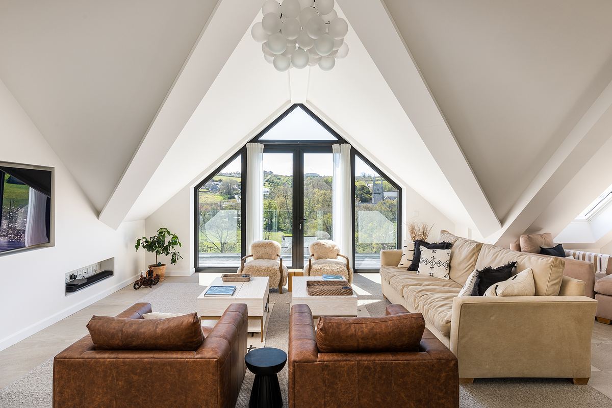 Spacious attic living room with triangular ceiling, large windows, beige sofas, brown armchairs, and a modern light fixture.