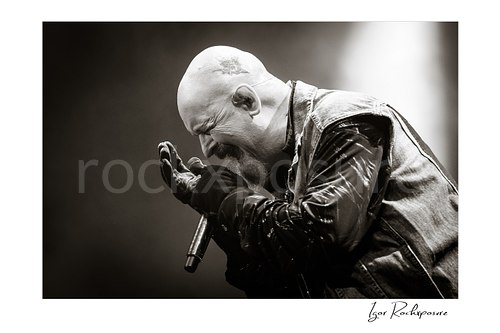 Horizontal black and white close-up image of Rob Halford of Judas Priest performing live on stage, holding a microphone and singing with an intense expression under dramatic lighting