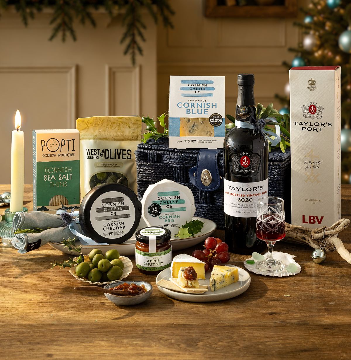 The contents of Cornish hamper with cheeses, crackers, olives, chutney, port wine, and glasses are arranged on a table with festive decorations and a lit candle in the background.