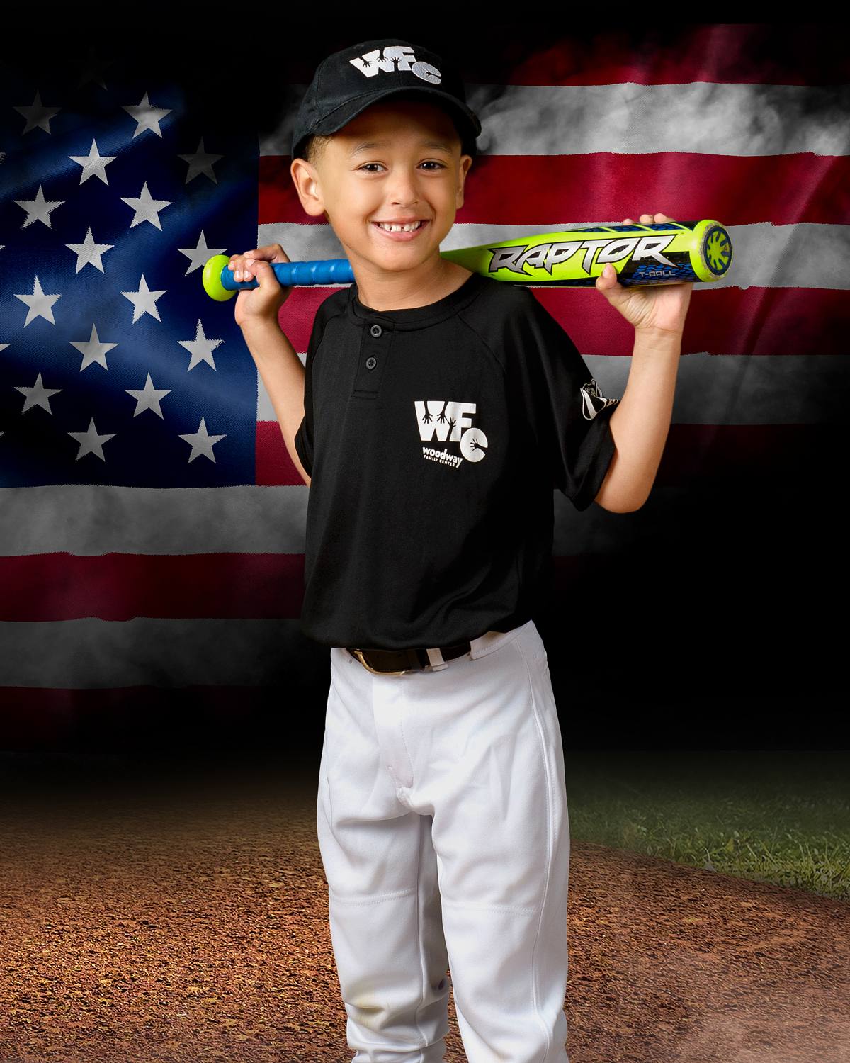 Smiling young t-ball baseball player holding a bat on his shoulders in front of a large American flag background on a baseball field. | ProClicks Photography