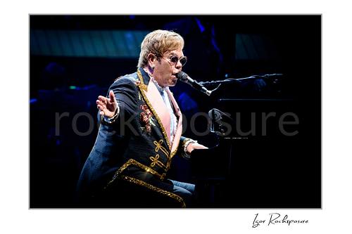 Horizontal color image of Elton John singing at the piano with one hand extended in a dark blue concert scene