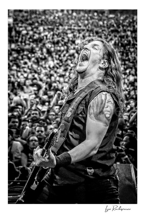 Vertical black and white image of Frank Bello of Anthrax performing live on stage, shouting while playing bass guitar with a large crowd visible in the background
