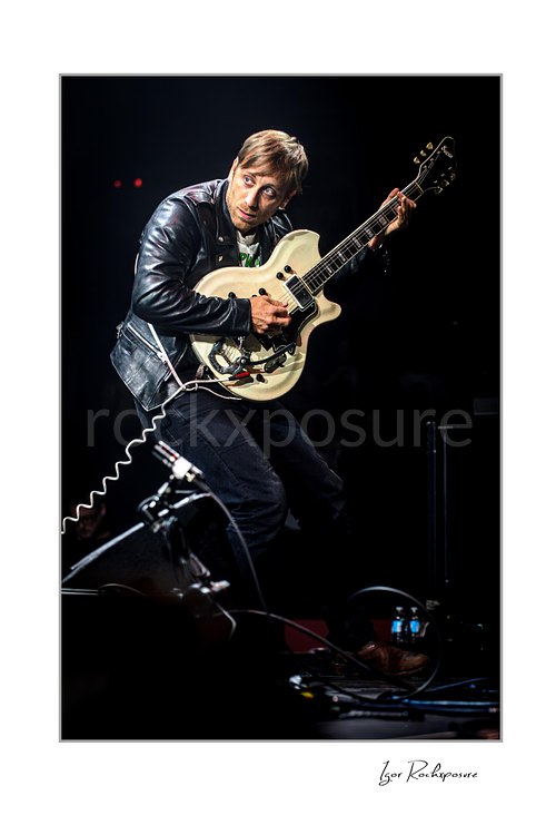 Vertical color image of Dan Auerbach of The Black Keys crouching with a white guitar in a dark concert scene