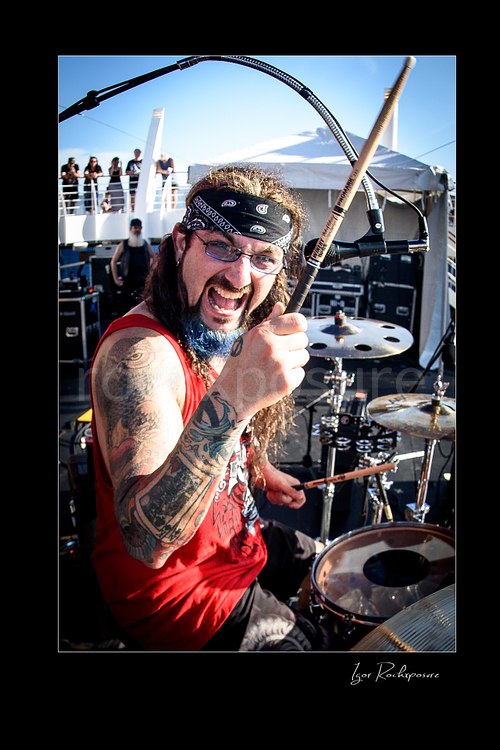 Vertical color image of Mike Portnoy at the drum kit grinning and holding a drumstick toward the camera in bright daylight