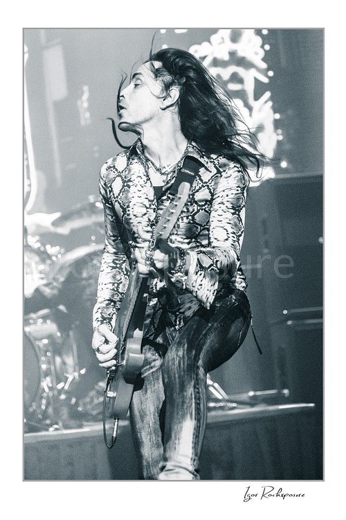 Vertical black and white image of Nuno Bettencourt performing live on stage with an electric guitar and windblown hair in dramatic motion