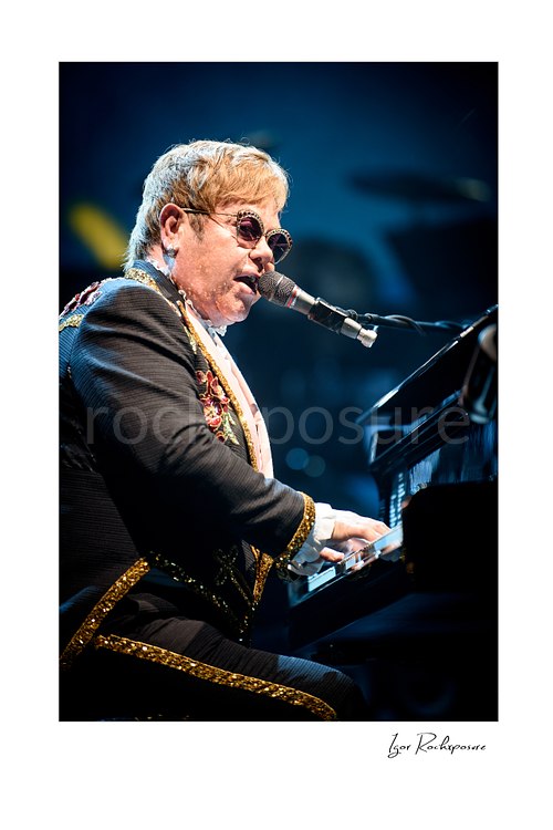 Vertical color image of Elton John singing at the piano in close-up under soft blue stage lighting