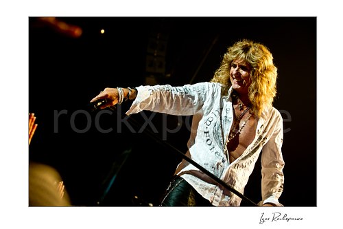 Horizontal color image of David Coverdale of Whitesnake performing live on stage, smiling while holding a microphone and mic stand under warm concert lighting