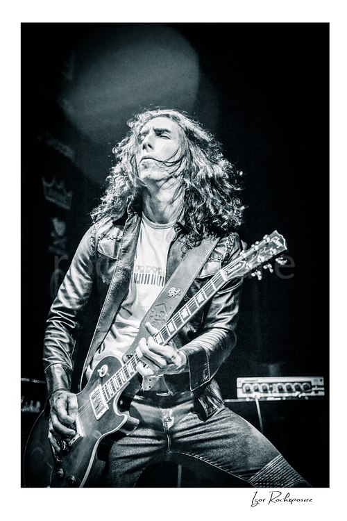 Vertical black and white image of Brent Woods playing guitar on stage with long hair and dramatic overhead light
