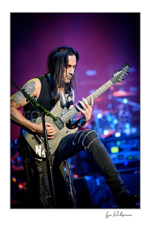 Vertical color image of Nuno Bettencourt performing live on stage with an electric guitar, one foot raised, under vivid purple and blue lighting