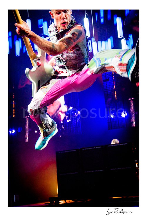 Flea of Red Hot Chili Peppers leaps with one leg extended, gripping a white bass above stage monitors under blue LED lights in colorful trousers.