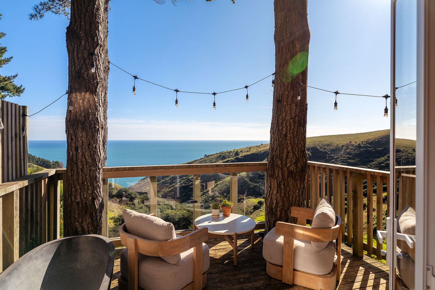 The outdoor balcony and seating area of the iconic Raglan Treehouse showcasing the expansive ocean view framed by mature trees, photographed by Flax Cove Studio