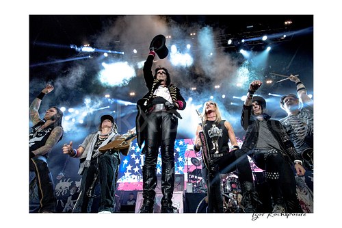Horizontal color image of Alice Cooper performing live on stage with the his Band, raised top hat, stage smoke, and bright concert lights