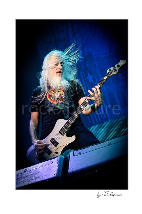 Vertical color image of John Campbell of Lamb of God playing bass on stage with flying white hair and blue concert light