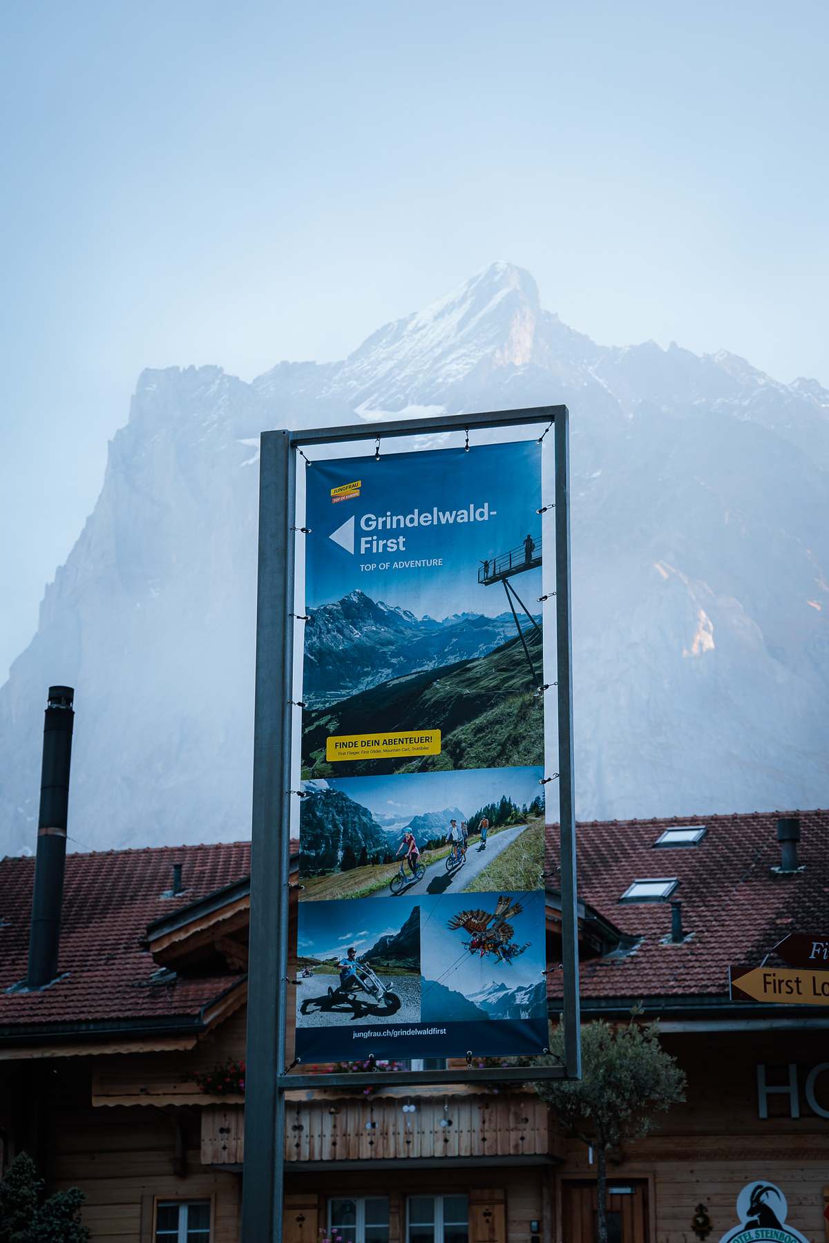 Werbebanner f&uuml;r Grindelwald-First mit Cliff Walk, First Flieger, Mountaincart und First Glider vor dem Hotel Steinbock und einem m&auml;chtigen Berggipfel im Dunst