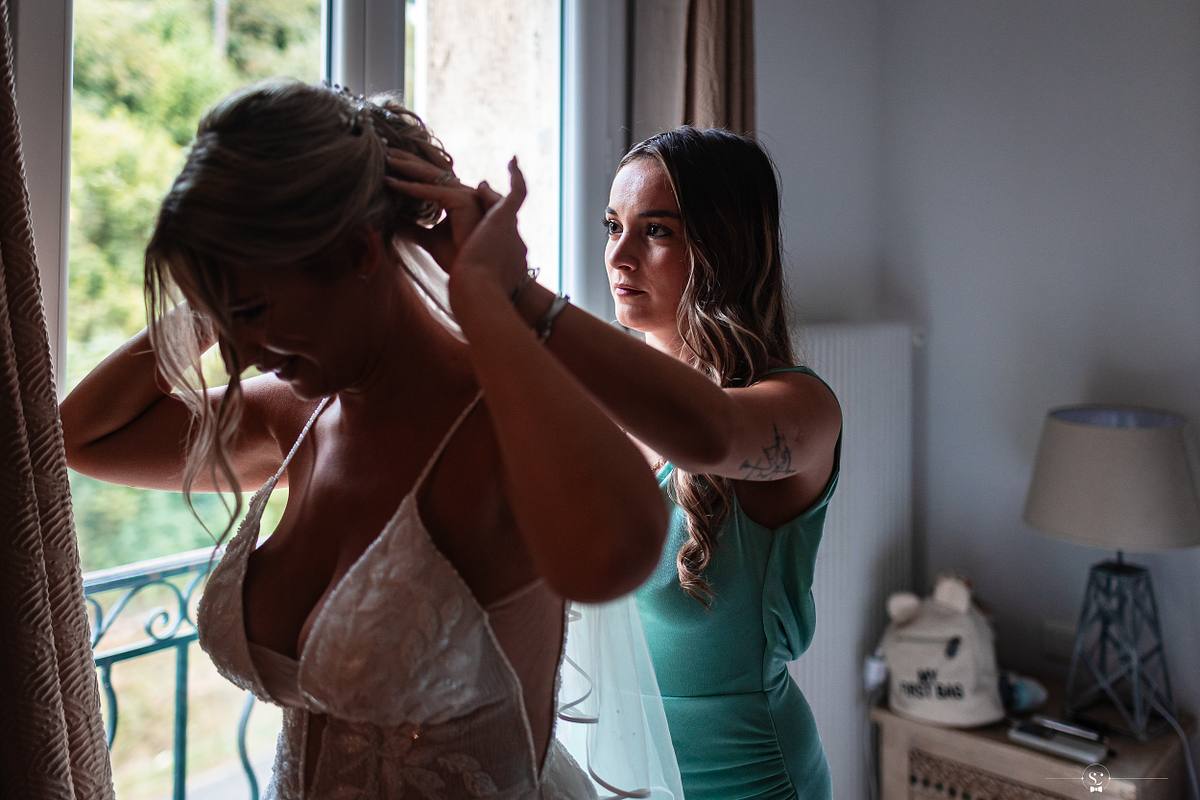 La t&eacute;moin en tenue de soir&eacute;e aide la mari&eacute;e &agrave; ajuster son voile dans une chambre lumineuse, soulignant le r&ocirc;le de la famille et des proches dans les moments pr&eacute;cieux avant la c&eacute;r&eacute;monie &agrave; N&icirc;mes