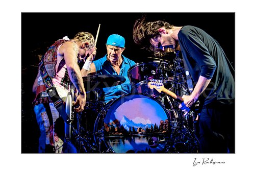 Flea and Josh Klinghoffer bend over their instruments on either side of Chad Smith, who raises a drumstick above purple toms and a landscape bass drum.