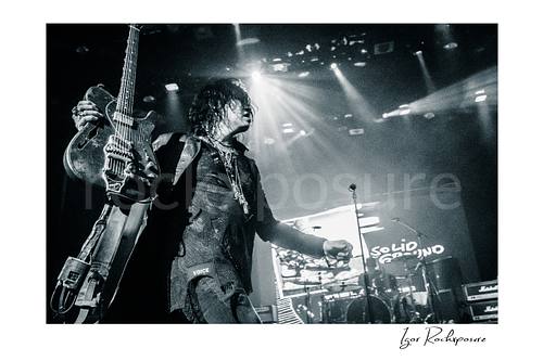Horizontal black and white image of Tom Keifer on stage holding his guitar upright in a dramatic live performance moment, with bright concert beams and the drum kit visible behind him