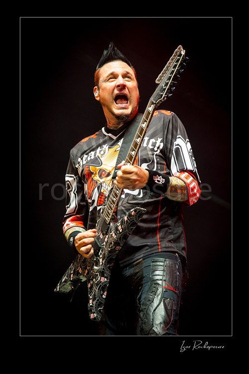 Vertical color image of Zoltan Bathory of Five Finger Death Punch playing guitar in a tight live performance shot under warm stage light