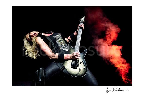 Horizontal color image of Nita Strauss performing live on stage with an electric guitar, leaning back with dramatic red stage smoke behind her
